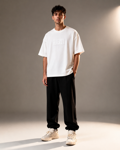 A man modeling a white short-sleeve t-shirt with a tonal rectangular logo on the chest, paired with black jogger pants and beige sneakers, set against a warm gray studio backdrop.