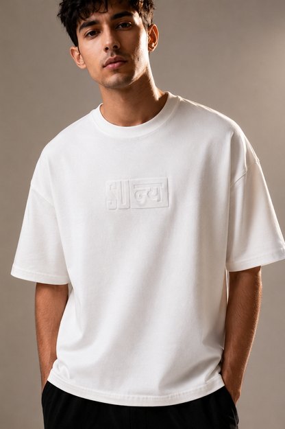 A man wearing a white short-sleeve crewneck t-shirt with a tonal embossed rectangular logo on the chest, paired with black pants, against a neutral studio background.