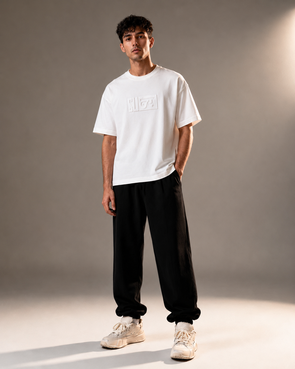 A man modeling a white short-sleeve t-shirt with a tonal rectangular logo on the chest, paired with black sweatpants and beige sneakers, set against a neutral gray studio background.