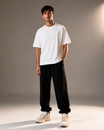 A man modeling a white short-sleeve t-shirt with a tonal rectangular logo on the chest, paired with black sweatpants and beige sneakers, set against a neutral gray studio background.
