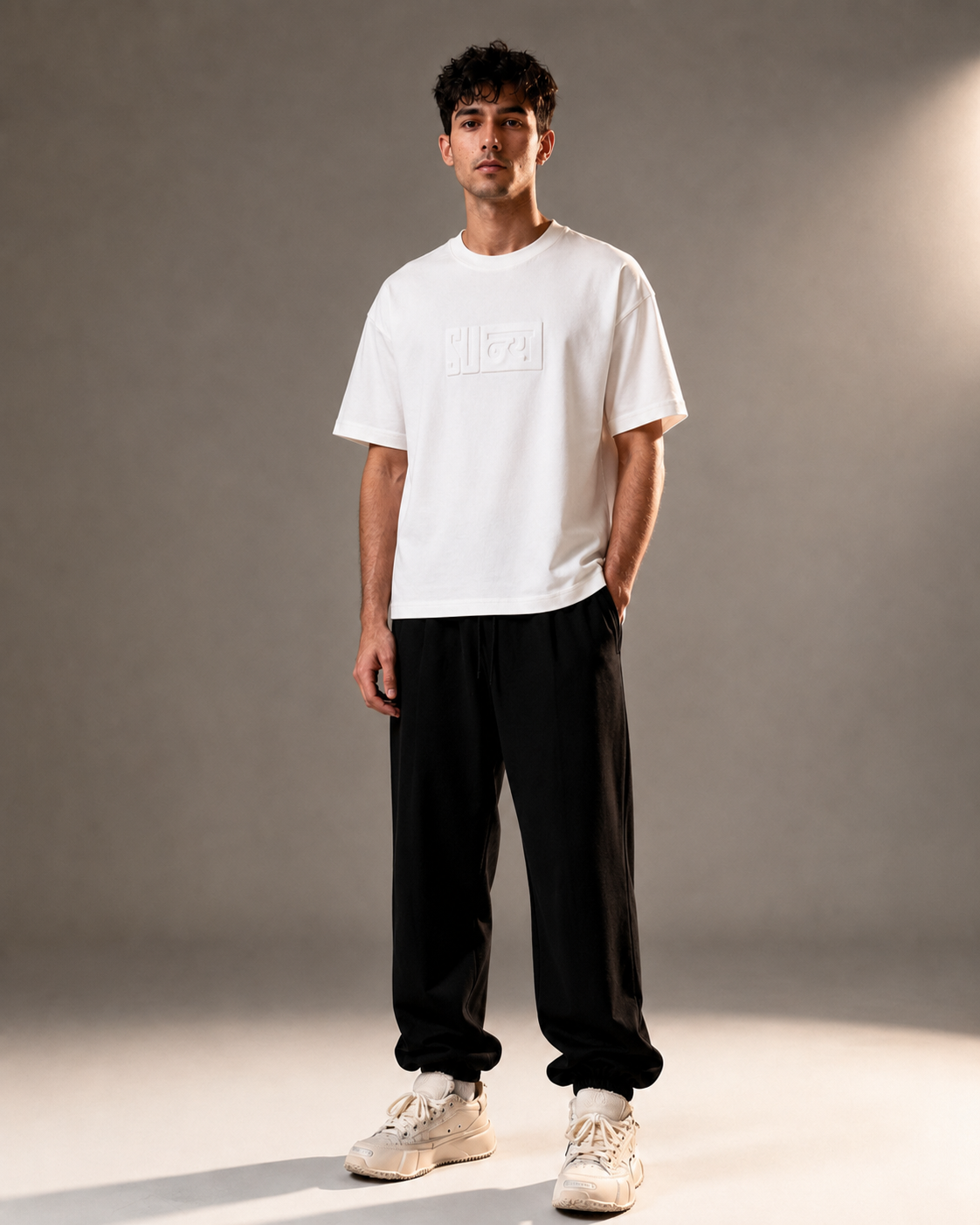 A man modeling a white short-sleeve t-shirt with a tonal rectangular logo on the chest, paired with black jogger pants and beige sneakers, standing against a gray studio backdrop.