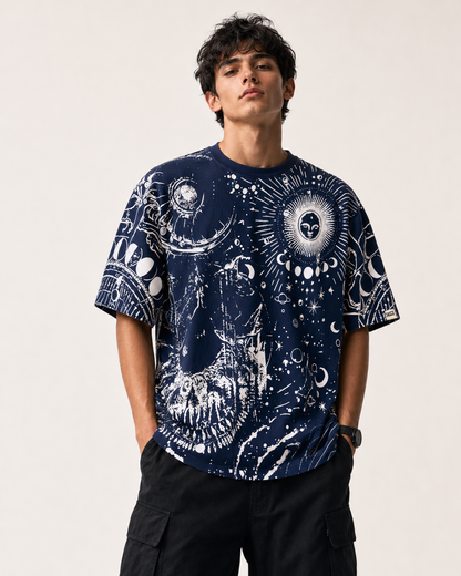 A man wearing a navy blue short-sleeve t-shirt with a white cosmic graphic print featuring skulls, aliens, and celestial patterns, paired with black cargo pants against a light beige background.