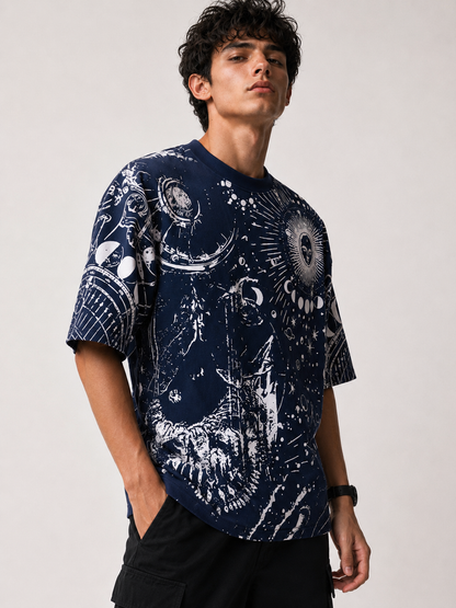 A man wearing a navy blue short-sleeve t-shirt with a white cosmic and celestial graphic print, paired with black cargo pants, photographed against a neutral light gray background.