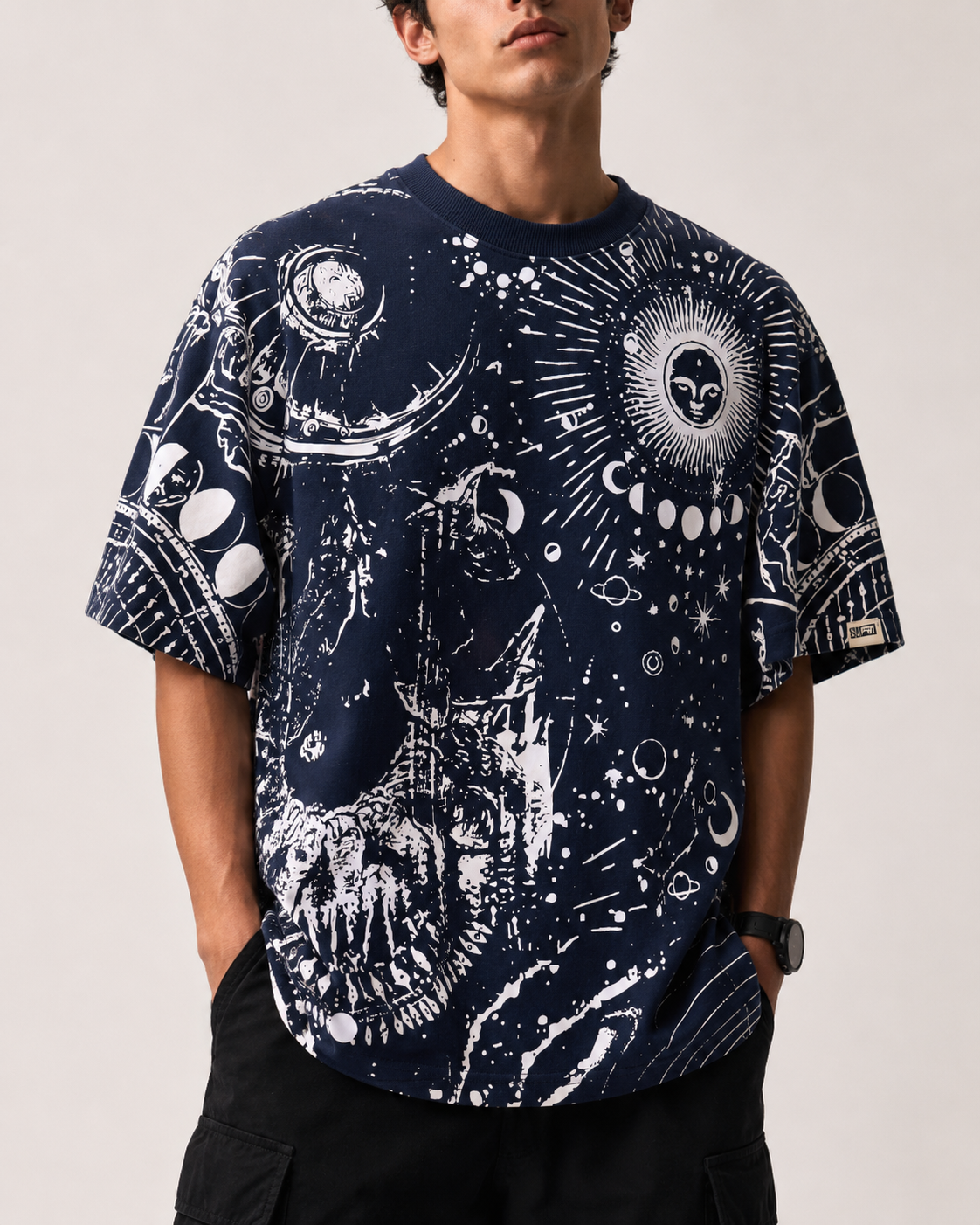 A man wearing a navy blue short-sleeve t-shirt with a white celestial and alien-themed graphic print, paired with black cargo pants against a light beige studio background.
