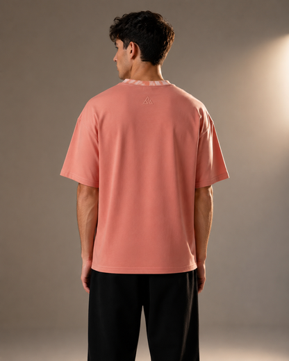 A man viewed from behind wearing a peach-pink short-sleeve t-shirt with a small embroidered logo on the upper back, paired with black pants, against a warm gray studio background.