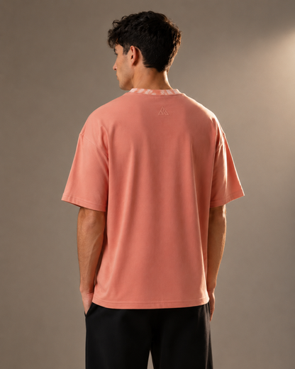 A man viewed from behind wearing a peach-colored short-sleeve t-shirt and black pants, standing against a neutral gray studio background.