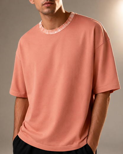 A man wearing an oversized short-sleeve t-shirt in a dusty peach color with a contrasting textured light pink ribbed crew neck collar, shown from the chin down against a neutral studio background.