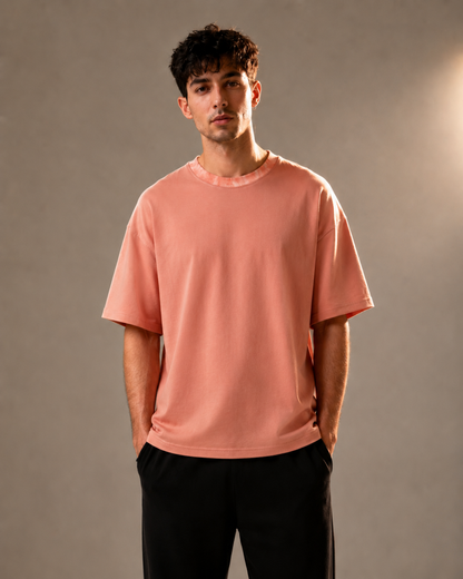 A man wearing a short-sleeve crewneck t-shirt in a dusty rose pink color, paired with black pants, standing against a neutral gray studio background.