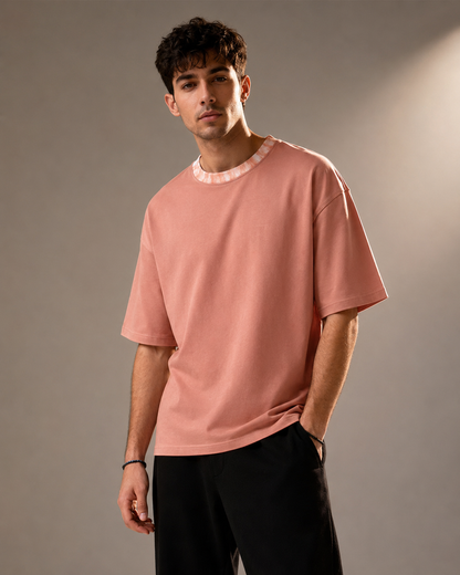 A man wearing a dusty pink short-sleeve t-shirt with a contrasting light pink patterned crew neck trim, paired with black pants, standing against a neutral gray studio background.